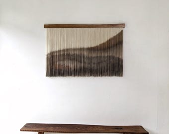 May include: A wall hanging composed of numerous vertical cream and brown yarn strands suspended from a horizontal wooden bar. Below the wall hanging sits a long, dark brown wooden bench with a natural live edge and four sturdy legs.