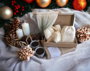 May include: A holiday-themed flat lay featuring a gift box with craft supplies, candles, and ornaments. The box contains yarn, a comb, and a drawstring bag. The scene is decorated with pinecones, beads, and two cups of coffee.