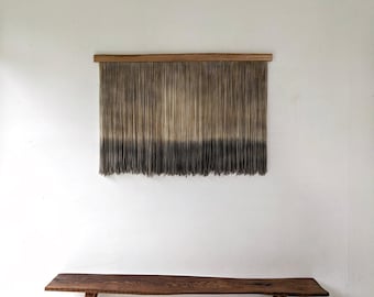 May include: A wall hanging featuring a horizontal wooden bar with numerous vertical hanging threads that transition in color from a light tan at the top to a dark charcoal gray at the bottom, positioned above a wooden bench with a live edge.