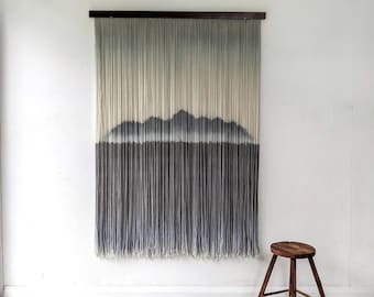 May include: A large wall hanging with a mountain design. The artwork features vertical strands in shades of gray and blue, creating a gradient effect. It is mounted on a dark wooden bar. A small wooden stool sits nearby.