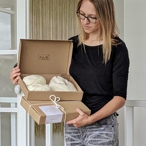 Puede incluir: Una persona sostiene dos cajas de cartón abiertas. La caja superior contiene dos madejas de hilo color crema y una pequeña bolsa con cordón. La caja inferior está cerrada y atada con hilo. El texto de la etiqueta dice "Hecho con amor y mucho hilo."