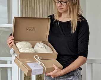 May include: A person holding two open cardboard boxes. The top box contains two skeins of cream-colored yarn and a small drawstring bag. The bottom box is closed and tied with twine. Text on the label reads "Crafted with love, and a whole lot of yarn."