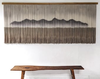 May include: A large, rectangular wall hanging with a mountain landscape design. The artwork features a wooden frame and long, vertical strands in shades of brown and gray. A wooden bench sits below the artwork.