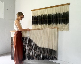May include: A woman stands in a room with two large wall hangings made of yarn. The hangings are white with black and brown stripes. The woman is holding one of the hangings in her hands.