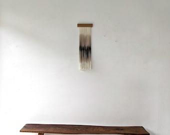 May include: A long wooden bench with a natural live edge and four legs, positioned below a wall hanging consisting of a horizontal wooden bar with vertical off-white fiber strands that transition into a dark charcoal gradient in the center.