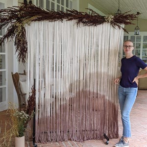 May include: A white and brown ombre macrame backdrop with a metal stand. The backdrop is made of yarn and is hanging from the stand. The backdrop is in front of a white wall and a person is standing in front of it.