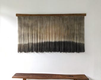 May include: A wall hanging featuring a horizontal wooden bar with numerous vertical beige strings that transition into a dark charcoal gray color at the bottom. Below the wall hanging is a wooden bench with a natural live edge finish.