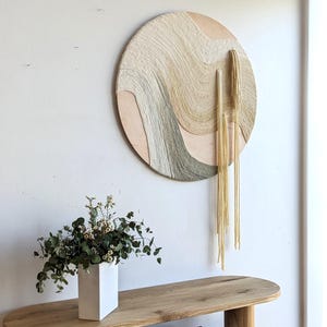 May include: A round wall hanging with a woven design in neutral tones of beige, gray, and gold. The artwork features a layered texture and hanging gold strands. A wooden console table below holds a white vase with greenery and books.