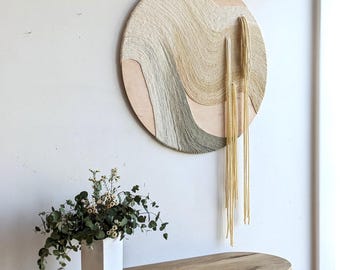 May include: A round wall hanging with a woven design in neutral tones of beige, gray, and gold. The artwork features a layered texture and hanging gold strands. A wooden console table below holds a white vase with greenery and books.
