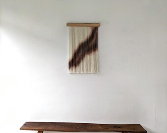 May include: A wall hanging featuring a series of vertical cream-colored fiber strands suspended from a horizontal wooden bar. A diagonal gradient of dark brown and black tones runs across the center of the fibers, creating a distinct color pattern.