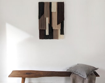 May include: A rectangular wall hanging with a textured design in shades of black, brown, and beige. Below, a wooden bench with a live edge supports a gray pillow and a fringed blanket. The scene is set against a white wall and a gray floor.