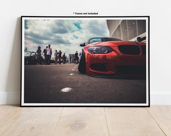 May include: A red sports car parked in a parking lot with a group of people walking in the background. The car has a black front bumper and black wheels.