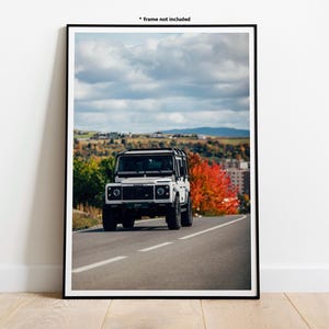 May include: Framed print of a white SUV on a road, set against autumn trees and a cloudy sky. The print is displayed on a wooden floor. The frame is black, and the text '* frame not included' is visible.