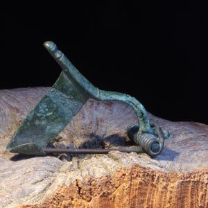 Puede incluir: Un broche antiguo de bronce, con un alfiler curvado y una placa angular decorativa. El broche tiene una pátina verde desgastada, que sugiere antigüedad. Descansa sobre una superficie de madera texturizada, sobre un fondo negro.