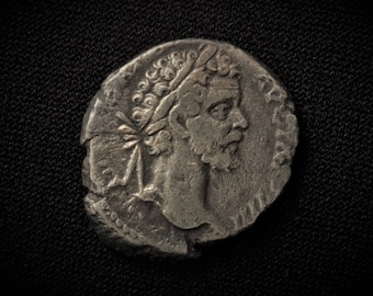 May include: A silver Roman coin with a portrait of a man with a laurel wreath on his head. The coin has a raised rim and is slightly worn.
