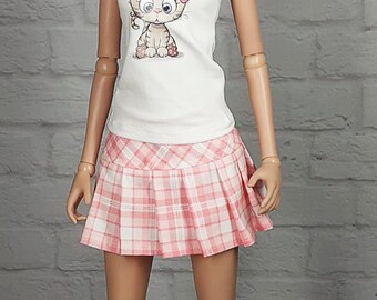 Könnte beinhalten: Eine Puppe trägt ein weißes T-Shirt mit einem Comic-Katzenmotiv und einen rosa-weiß karierten Rock. Die Puppe hat blonde Haare und trägt weiße Turnschuhe.