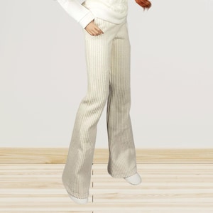 Könnte beinhalten: Eine Modepuppe mit langen, welligen, rotbraunen Haaren, die einen weißen Pullover mit strukturiertem Muster, ein weißes Langarmshirt und eine beige gestreifte Hose trägt. Die Puppe trägt auch weiße Schuhe. Die Puppe steht vor einem neutralen Hintergrund.