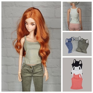 Op de afbeelding: Een pop draagt een groene tanktop en een corduroy broek. Extra afbeeldingen tonen de tanktop in verschillende kleuren: blauw, grijs en zwart. De pop heeft lang, rood haar en poseert voor een witte bakstenen muur.