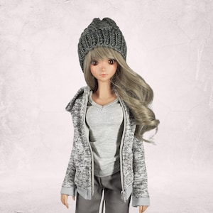 Könnte beinhalten: Eine graue Strickmütze mit einem grau-weißen Kapuzenpullover und einer grauen Hose. Die Puppe hat langes blondes Haar.