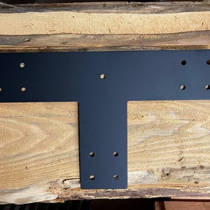May include: Black metal T-shaped bracket with multiple holes for attaching to wood. The bracket is lying on a wooden surface.
