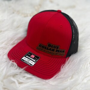 Op de afbeelding: Rode en zwarte trucker cap met een bruin leren patch waarop "BLUE COLLAR MAN with a crazy of lady" staat. De pet heeft een Richardson-logo en de tekst "ORIGINAL 112 TRUCKER ADJUSTABLE".