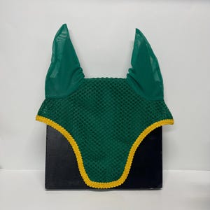 May include: A dark green equestrian fly bonnet with a yellow trim. The bonnet has mesh ear coverings and a crocheted body. The item is designed to protect horses from insects and is likely used during riding or competitions.