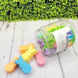 May include: A clear jar filled with colorful bunny-shaped soaps. The jar is tipped over, spilling out some of the soaps. The soaps are in various colors, including pink, blue, yellow, and orange. The jar has a label that says "Spring Bunny Soaps" and "Handmade with Love".