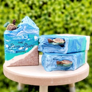 May include: Three rectangular bars of handmade soap featuring a blue and white swirled ocean design with a sandy brown base. Each bar is topped with a small, detailed brown and green sea turtle figure. The front bar displays the text THE Soap Swirl SHOPPE.