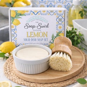 May include: A solid dish soap set in a white box with lemon and tile patterns, labeled The Soap Swirl Shoppe Lemon Solid Dish Soap Set, net weight 6 oz. Includes a round white ceramic dish containing solid soap and a wooden scrub brush.