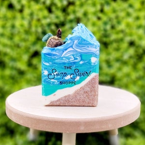 May include: A rectangular bar of soap featuring a beach scene with a sandy bottom, a white foam line, and blue ocean waves. A small turtle figure sits on top of the soap. The text "THE Soap Swirl SHOPPE" is printed in black on the front.