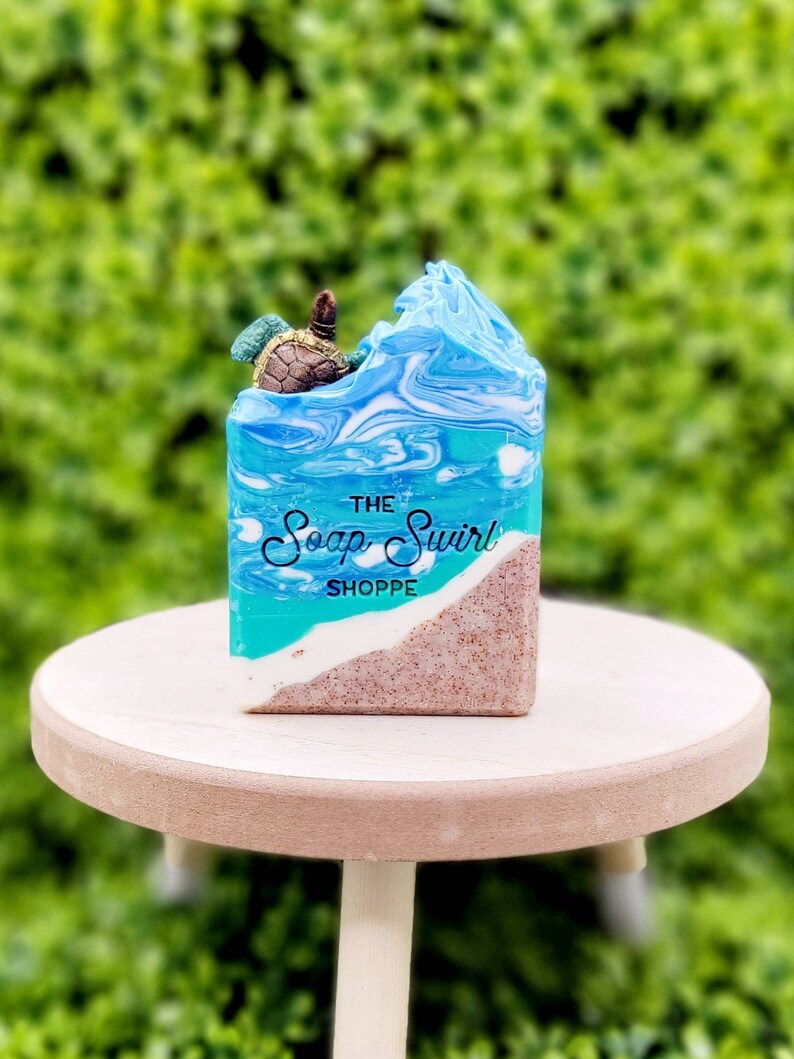 May include: A rectangular bar of soap featuring a beach scene with a sandy bottom, a white foam line, and blue ocean waves. A small turtle figure sits on top of the soap. The text "THE Soap Swirl SHOPPE" is printed in black on the front.