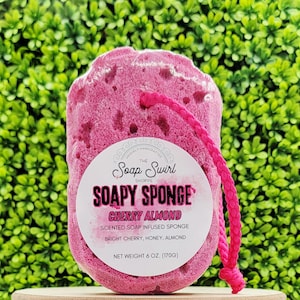 May include: A pink, textured, cherry almond scented soap-infused sponge with a pink cord loop. The label reads "SOAPY SPONGE" and "CHERRY ALMOND". Net weight is 6 oz (170g).