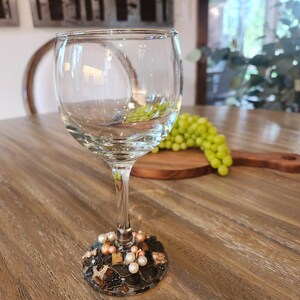 May include: A clear wine glass with a decorative base featuring pearls, copper-colored beads, and dark brown accents. The glass is empty, and a bunch of green grapes rests on a wooden cutting board in the background.