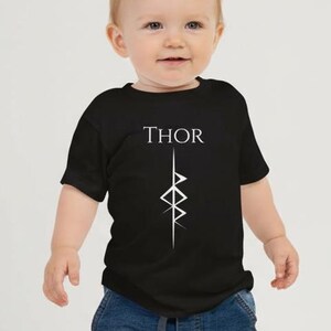 以下が含まれることがあります： 白いグラフィックデザインの上に、白い文字で「THOR」と書かれた黒いTシャツ。 青いジーンズと白いスニーカーを履いた赤ちゃんが着ています。