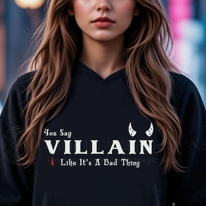 Young woman with long brown hair in an urban setting wearing a black villain hoodie that says You Say Villain Like It's A Bad Thing with devil horns created by graphic apparel company Midnight Gypsy Designs as part of their bookish merch clothing line.