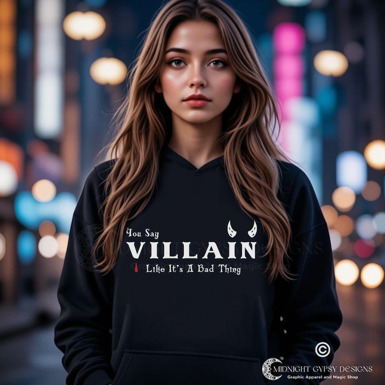 Young woman with long brown hair in an urban setting wearing a black villain hoodie that says You Say Villain Like It's A Bad Thing with devil horns created by graphic apparel company Midnight Gypsy Designs as part of their bookish merch clothing line.