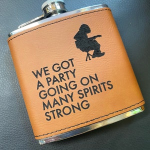 May include: A brown leather-wrapped flask with a silver metal rim and cap. The flask has a black silhouette of a person sitting in a chair and the text "WE GOT A PARTY GOING ON MANY SPIRITS STRONG".
