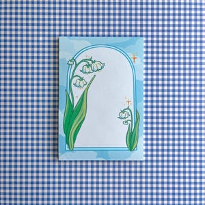 Puede incluir: Un bloc de notas con temática floral con una ilustración de lirios de los valles. El diseño incluye un borde arqueado azul claro con un fondo similar al cielo. El bloc de notas está sobre un patrón de cuadros azules y blancos.