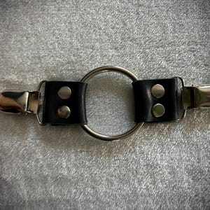 以下が含まれることがあります： 中央に大きなシルバーリングが付いた、黒革と金属製のアクセサリー。シルバーの金属クリップが付いた2つの黒革片がリングに取り付けられています。各革片には2つのシルバーリベットが付いています。