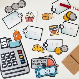 K&ouml;nnte beinhalten: Eine Kasse mit einer Kreditkarte, Spielgeld und Preisschildern f&uuml;r einen Spielwarenladen. Die Preisschilder beinhalten Artikel wie einen Cupcake, Brot, M&uuml;sli, K&auml;se und Orangensaft. Die Preise sind in Britischen Pfund.