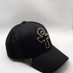 May include: Black baseball cap with a gold sequined ankh symbol on the front. The brim has a row of gold sequins. The cap is made of a textured fabric and has a rounded top.