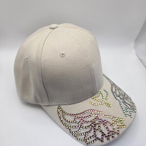 May include: A beige baseball cap with a curved peak. The peak is adorned with colourful rhinestones arranged in a wing design. The cap has a button on top and is made of a textured fabric.