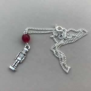 Può includere: Una collana color argento con un piccolo ciondolo a forma di schiaccianoci e una perla rossa. La catena è delicata e poggia su uno sfondo neutro. Il ciondolo schiaccianoci è alto circa 2,5 cm.