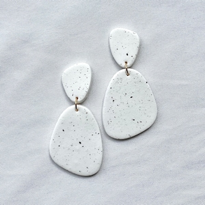 May include: A pair of white, speckled earrings with a gold-colored hook. The earrings have an organic, layered shape, with a smaller top piece connected to a larger bottom piece. The speckled design features small black and gray dots.