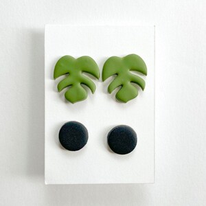 May include: A white card displays two pairs of earrings. One pair features green monstera leaf designs, the other pair is black, circular studs. The earrings are likely made from polymer clay or a similar material. The leaf earrings are a light green.