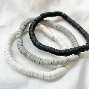 May include: Three beaded bracelets in a grayscale palette. The bracelets are made of small, flat, disc-shaped beads in white, gray, and black. The bracelets are arranged on a white, textured surface.
