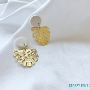 May include: A pair of gold-colored earrings featuring a monstera leaf design. Each earring has a small, round, off-white stud at the top. The earrings are hanging on a white fabric background. The text "Starry Days Jewelry" is visible in the bottom right corner.
