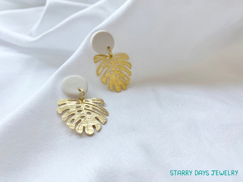 May include: A pair of gold-colored earrings featuring a monstera leaf design. Each earring has a small, round, off-white stud at the top. The earrings are hanging on a white fabric background. The text "Starry Days Jewelry" is visible in the bottom right corner.