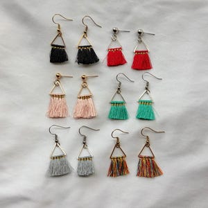 May include: A collection of handmade tassel earrings in various colors, including black, red, pink, teal, gray, and multicolored. Each earring features a triangular metal frame and a fringe of soft tassels. The earrings are displayed on a white surface.