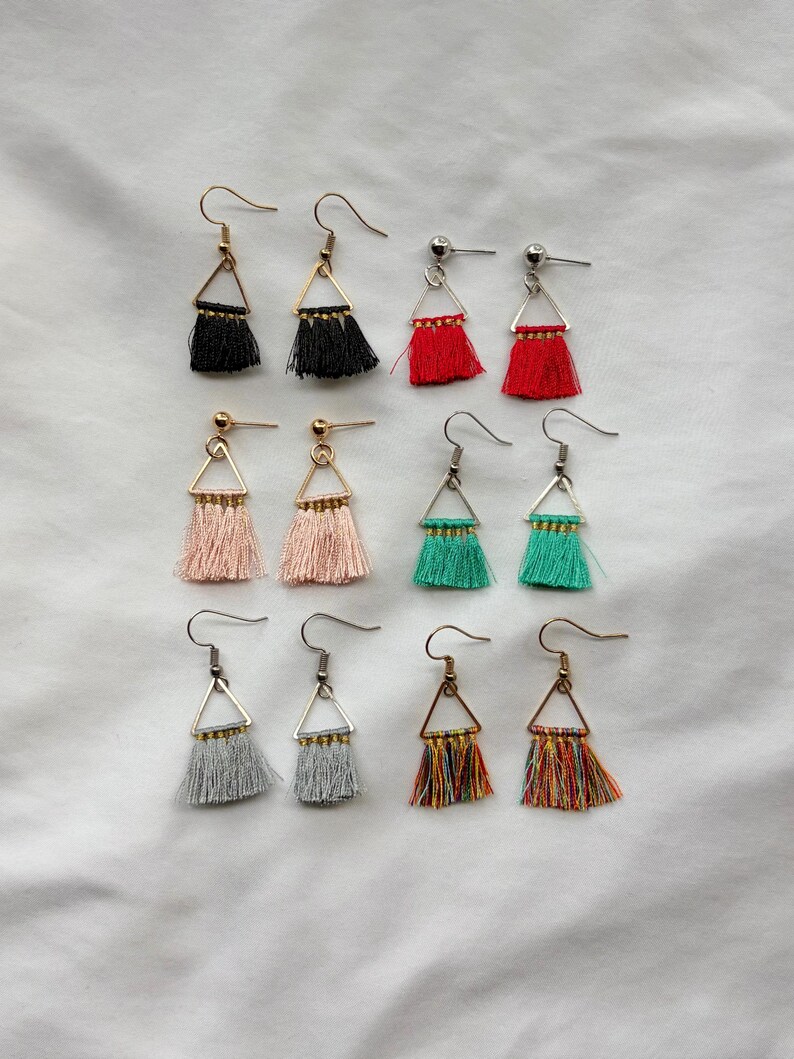 May include: A collection of handmade tassel earrings in various colors, including black, red, pink, teal, gray, and multicolored. Each earring features a triangular metal frame and a fringe of soft tassels. The earrings are displayed on a white surface.